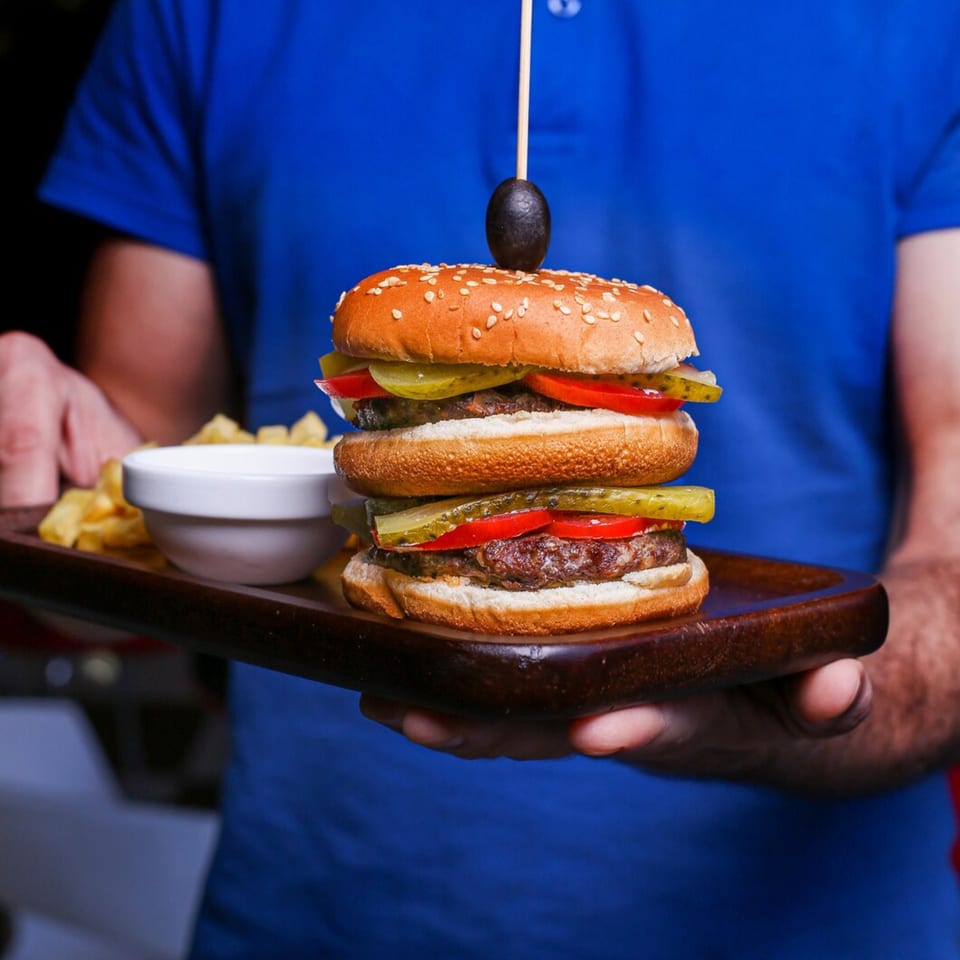 A server in a blue shirt holds a wooden tray with a double Burger Shack burger, fries, and dip.