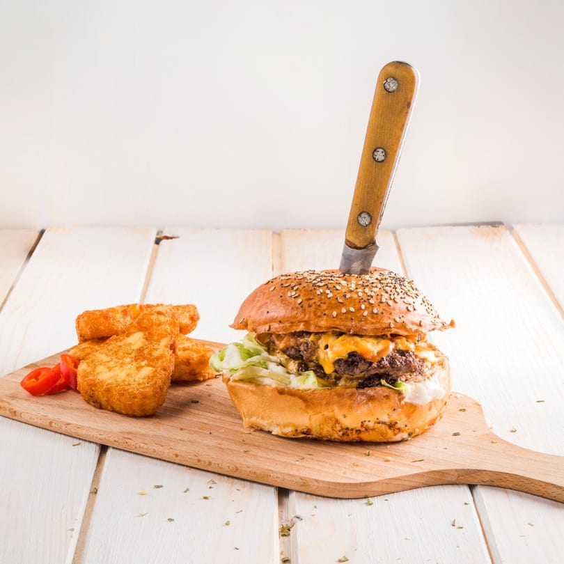 A Burger Shack burger with melted cheese and lettuce, skewered by a knife, served with hash browns.