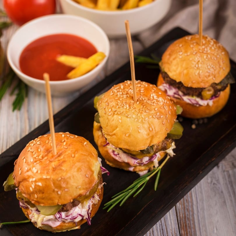 Three Burger Shack sliders with pickles and coleslaw on a dark wooden board, with ketchup and fries.