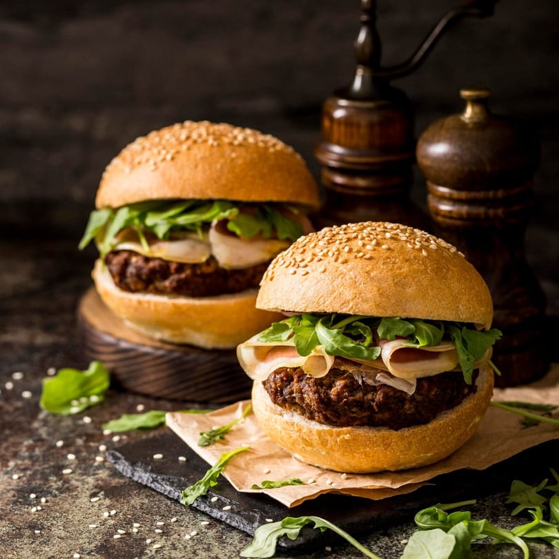 Two Burger Shack burgers with arugula, cheese, and sesame seed buns on a dark surface.