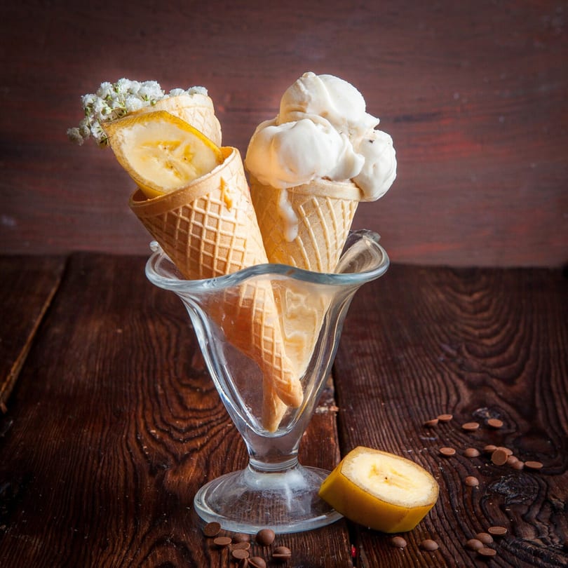 Vanilla ice cream cone and banana slices in waffle cones, in a glass dish on a wooden table.